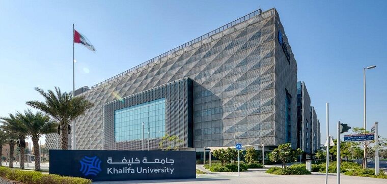 A huge grey building of Khalifa university and sidewalk, palm trees and gardens.